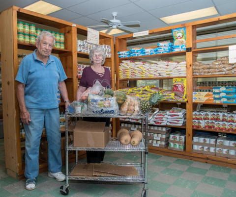 Two aduls age 60+, one man and one woman stand looking proud with lots of shelves of behind them full of boxed and canned food items. 