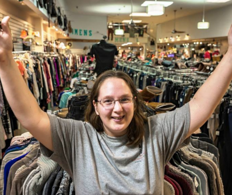 A young woman approximately in her early to mid 20's stands infront of racks of clothes. She has long light brown hair and glasses. She stands with her arms triumphantly spread over her head with a smile on her face. 