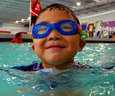 All boy approximate 5 years old smiles for the camera. He is in an indoor pool with blue swim goggles on. He has wet dark hair.