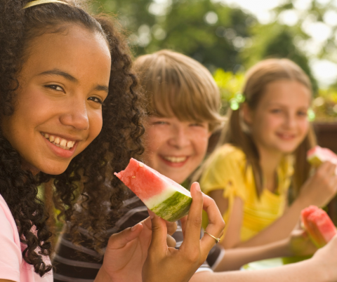 3 kids approximate 8-10 years old, 1 black girl wearing pink with long curls, one little boy white, white with shaggy, sandy colored hair, one girl white with sandy colored hair in pigtails, all looking at the camera and smiling with watermelon in their hands.